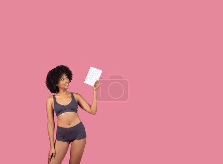 Smiling, active young black woman in fitness attire confidently holding up menstrual cycle tracker calendar against soft pink background, copy space