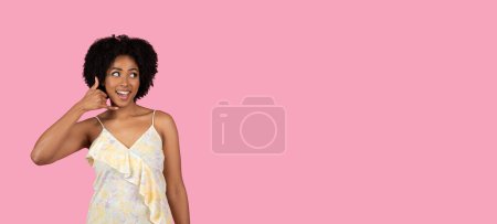 Photo for Vivacious smiling millennial African-American woman with a playful expression making a call me gesture, dressed in a summery floral frock against a pink backdrop, studio, panorama - Royalty Free Image