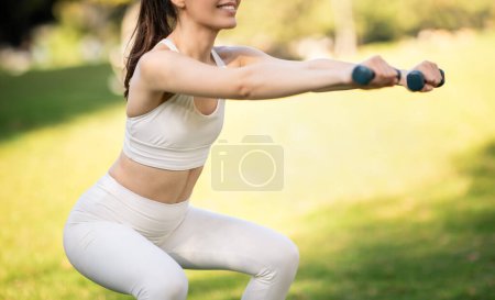 Photo for Focused young caucasian woman athlete in stylish sportswear performing a squat with hand weights in a sunlit park, embodying health and fitness, cropped. Sport, fitness, body care - Royalty Free Image