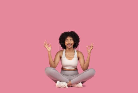 Foto de Mujer afroamericana sonriente en sujetador deportivo blanco y leggins grises sentados con piernas cruzadas en pose meditativa con los ojos cerrados sobre fondo rosa, espacio libre - Imagen libre de derechos
