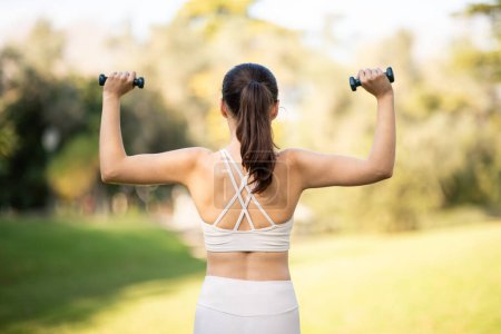 Photo for Rear view of a fit caucasian millennial woman exercising with dumbbells in a sunny park, showcasing her strength and healthy lifestyle, back. Power, sport outdoors, fit and energy - Royalty Free Image