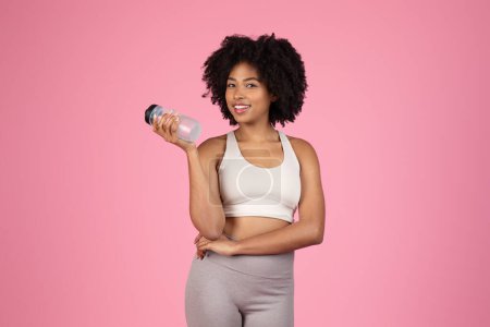 Téléchargez les photos : Joyeux jeune femme noire aux cheveux bouclés, vêtue de soutien-gorge de sport blanc et leggings gris, tenant bouteille d'eau, avec pose confiante sur fond rose - en image libre de droit