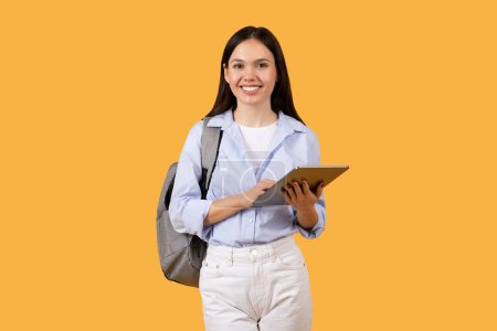 Photo for Cheerful female student exudes confidence and readiness for learning, standing with tablet, embodying modern educations blend of mobility and technology, yellow backdrop - Royalty Free Image