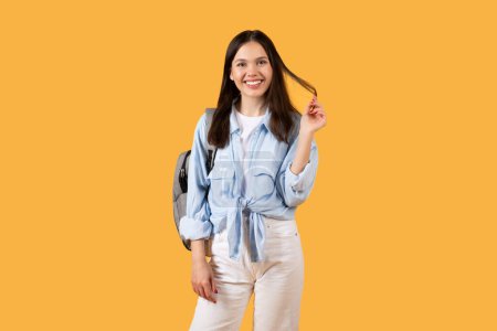 Cheerful lady student in casual attire wearing backpack and smiling at camera, exuding the optimism and readiness for her academic pursuits, yellow backdrop