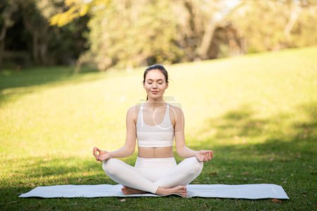 Photo for A focused caucasian young woman athlete in a white sports bra and leggings sits cross-legged on a yoga mat, practicing meditation in a sunny, green park, outside, full length - Royalty Free Image