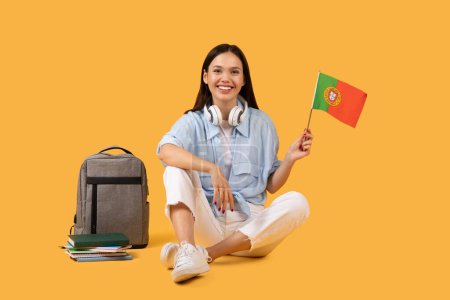 Photo for Content female student, dressed casually, sits cross-legged with her laptop, books and Portuguese flag, headphones around her neck, signifying blend of technology, education and cultural pride - Royalty Free Image