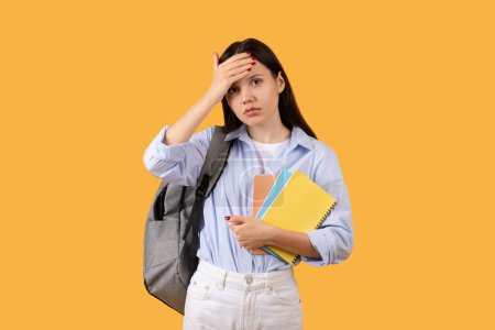 Concerned lady student with backpack and colorful notebooks stands against yellow background, touching forehead in gesture of worry, stress or confusion