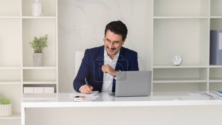 Photo for Laughing middle aged european businessman in a navy suit excitedly punching the air in triumph while working at his laptop in a clean, organized white office space, panorama - Royalty Free Image