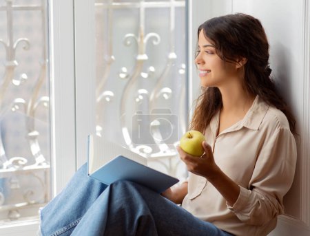 Photo for Smiling young woman enjoying healthy snack and reading book by the window at home, happy millennial female relaxing on floor in room, looking out at city view, relaxing and daydreaming, copy space - Royalty Free Image