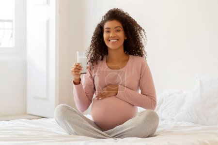 Téléchargez les photos : Femme enceinte noire souriante assise sur le lit et tenant un verre de lait, heureuse femme enceinte afro-américaine choisissant une boisson riche en calcium pendant la grossesse, recommandant une alimentation nutritive - en image libre de droit
