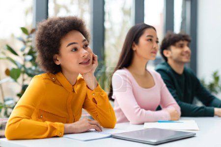Photo for Young black woman with curly hair looks contemplative, resting her chin on her hand, while her focused classmates listening in the background - Royalty Free Image