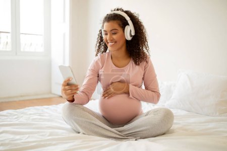 Foto de Mujer embarazada negro feliz usando auriculares usando teléfono inteligente en casa, afroamericana embarazada madre disfrutando de la selección de música en su teléfono mientras está sentado en la cama en el dormitorio, espacio para copiar - Imagen libre de derechos