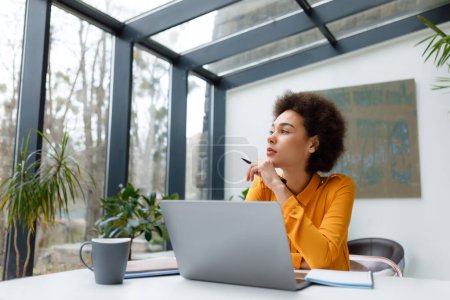 Photo for Contemplative young black woman with stylish afro hairstyle gazes out window while working at her laptop in bright, naturally lit modern office space - Royalty Free Image