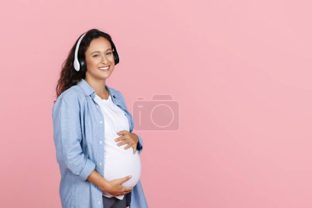 Téléchargez les photos : Une jeune femme enceinte positive écoutant de la musique, embrassant son ventre et souriant à la caméra, utilisant un casque sans fil, un fond de studio rose, un espace de copie. Impact de la musique sur le foetus - en image libre de droit