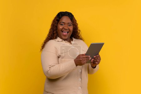 Téléchargez les photos : Happy chubby aux cheveux longs afro-américaine dame tenant tablette numérique et s'exclame, vérifier belle nouvelle application mobile, isolé sur fond jaune studio - en image libre de droit