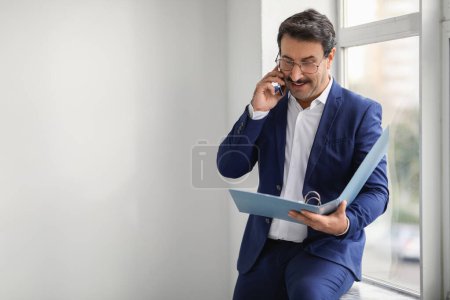 Photo for Corporate middle aged european businessman in a blue suit engaging in a phone conversation while reviewing a file near a bright window, displaying focus and efficiency in office - Royalty Free Image