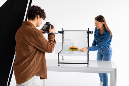 Foto de Fotógrafo experto en comida con chaqueta marrón enfoca intensamente su cámara en una hamburguesa de estilo perfecto, con un asistente ajustando la presentación en la mesa blanca - Imagen libre de derechos