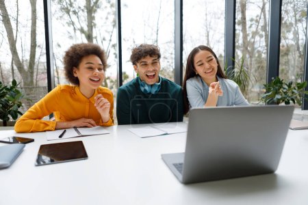 Foto de Tres estudiantes alegres diversos en ropa casual se absorben y ríen mientras miran la pantalla del ordenador portátil en el espacio de estudio brillante y moderno con vista a la ventana - Imagen libre de derechos