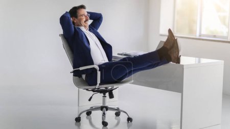 Téléchargez les photos : Homme d'affaires européen d'âge moyen, heureux et détendu, le sourire rayonnant, appuyé sur sa chaise, les jambes sur le bureau, profitant d'un moment de succès ou d'une pause dans un bureau lumineux - en image libre de droit