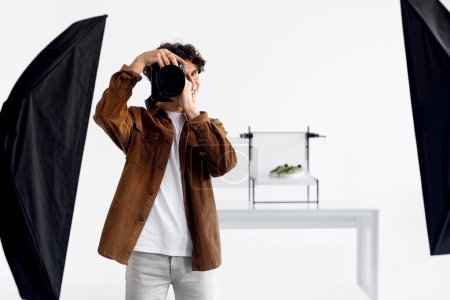 Photo for Professional male photographer in corduroy jacket focusing his DSLR camera, preparing to take photograph in well-lit studio with soft boxes, free space - Royalty Free Image