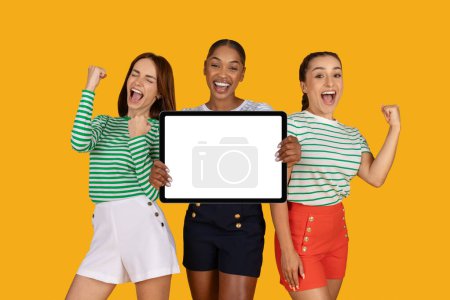 Photo for Overjoyed young multiethnic ladies girlfriends showing digital tablet with white blank screen mockup copy space gesturing and exclaiming, celebrating beginning of season sale, yellow background - Royalty Free Image