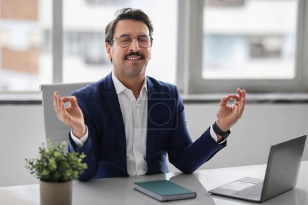 Zufriedener europäischer Geschäftsmann mittleren Alters im blauen Anzug meditiert an seinem Schreibtisch mit geschlossenen Augen und sucht inmitten eines geschäftigen Arbeitstages Ruhe. Arbeit, Geschäftspause