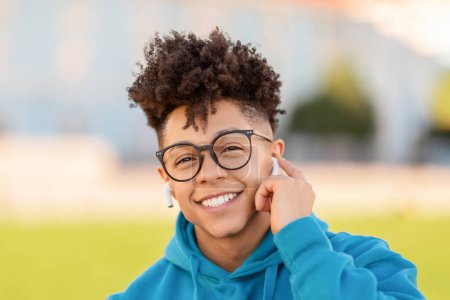 Téléchargez les photos : Portrait d'un jeune étudiant noir heureux avec des écouteurs sans fil souriant à la caméra, écoutant de la musique ou un podcast éducatif en ligne, passant du temps en plein air dans le parc du campus universitaire - en image libre de droit