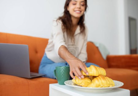 Foto de Mujer sonriente que busca croissants en el plato junto a su computadora portátil, mujer joven feliz tomando pastelería sabrosa, teniendo descanso mientras trabaja con la computadora en la sala de estar en casa, enfoque selectivo - Imagen libre de derechos