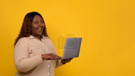 Téléchargez les photos : Femme afro-américaine millénaire potelée positive avec de longs cheveux bouclés portant une chemise beige à l'aide d'un ordinateur portable, tapant sur le clavier de l'ordinateur, regardant l'espace de copie, fond de studio jaune, panorama - en image libre de droit