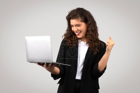 Téléchargez les photos : Femme d'affaires joyeuse avec les cheveux bouclés regardant l'ordinateur portable et levant son poing dans le geste victorieux, symbolisant le succès et la réalisation, fond gris - en image libre de droit