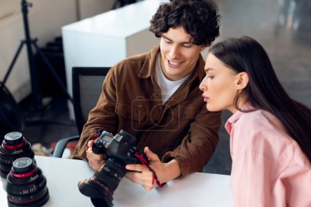 Foto de Joven fotógrafo masculino de pelo rizado muestra la pantalla de la cámara a una modelo femenina atenta en camisa rosa, colaborando durante la sesión de fotos del estudio - Imagen libre de derechos