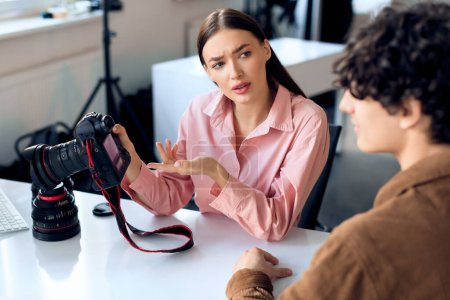 Photo for Female photographer discusses photographs with concerned male model, both reviewing camera shots, sitting at table in bright studio setting - Royalty Free Image