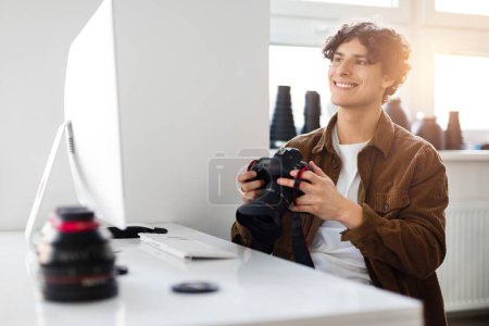 Foto de Alegre fotógrafo masculino con chaqueta de pana sonríe mientras revisa las fotos en su cámara réflex digital, sentado en un estudio bien iluminado con equipo de fotografía alrededor - Imagen libre de derechos