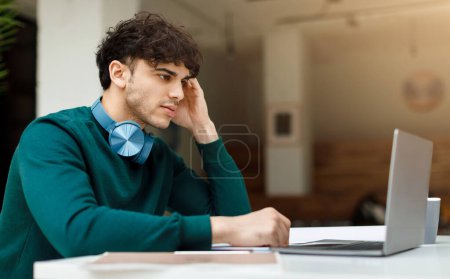 Photo for Thoughtful man student with headphones around his neck is studying or working on laptop, with hand to his temple, in bright office or classroom space - Royalty Free Image