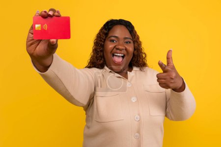 Photo for Cool chubby millennial african american woman with long curly hair pointing at red plastic credit card in her hand, recommending easy banking, isolated on yellow background - Royalty Free Image