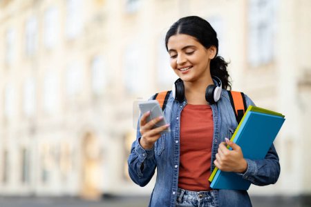 Photo for Communication concept. Cheerful indian woman student with wireless headphones and backpack texting her friends while walking by street university campus, carrying textbooks, using smartphone - Royalty Free Image