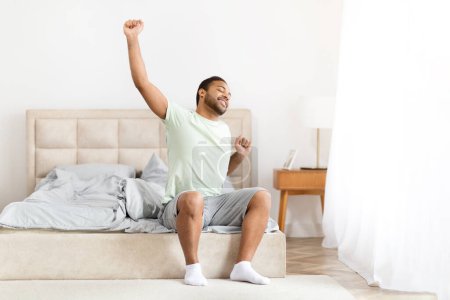 Photo for Happy well-rested millennial african american man wearing pajamas stretching his body while sitting in bed and looking through window, enjoying beginning of new day, sun flare, copy space - Royalty Free Image