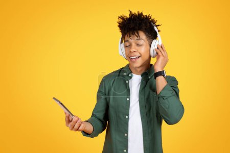 Téléchargez les photos : Joyeux jeune homme noir avec coiffure élégante écoutant de la musique isolée sur fond de studio jaune, en utilisant une application mobile sur smartphone et écouteurs sans fil, espace de copie - en image libre de droit