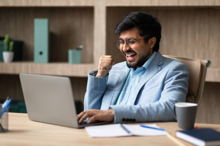 Téléchargez les photos : Réussite des affaires en ligne. Homme d'affaires indien excité à l'ordinateur portable célébrant la victoire dans l'intérieur du bureau, Gesturing Yes With Excitation lecture de grandes nouvelles Internet sur l'ordinateur au travail - en image libre de droit