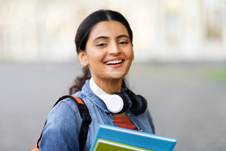 Téléchargez les photos : Portrait d'une belle étudiante arabe souriante avec écouteurs sans fil, sac à dos et livres à la main posant à l'extérieur, allant à l'université, espace de copie - en image libre de droit