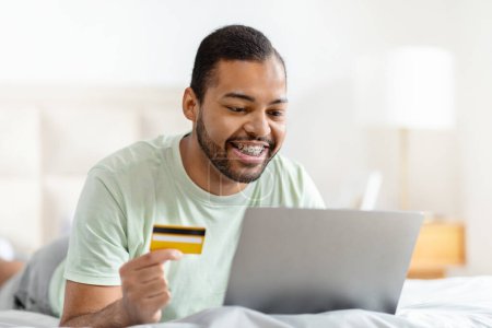 Relaxed bearded millennial african american man wearing pajamas lying on bed at home, using computer laptop and credit card, banking or shopping while resting at weekend, copy space