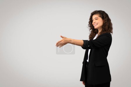 Foto de Mujer de negocios de pelo rizado en blazer negro extiende su mano para el apretón de manos, sonriendo de manera acogedora contra fondo gris neutro, espacio libre - Imagen libre de derechos