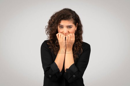 Photo for Worried young businesswoman in suit biting her nails, gesture of nervousness or stress, with concerned expression against grey background - Royalty Free Image