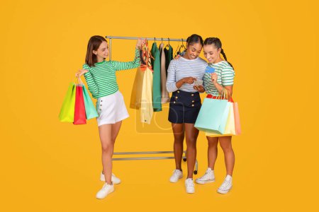 Photo for Three young multiethnic women girlfriends shopping together on yellow background, standing next to rack full of fashionable clothes, checking nice outfit, using smartphone and credit card - Royalty Free Image
