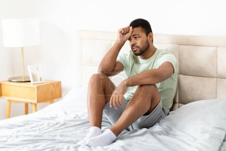 Photo for Unhappy handsome young african american man wearing pajamas homewear sitting on bed at home, suffering from depression, loneliness or anxiety, copy space. Permacrisis, midlife crisis - Royalty Free Image