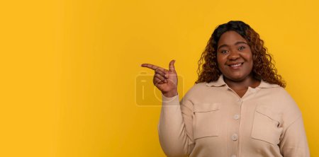 Photo for Positive curly chubby millennial black lady wearing beige shirt pointing at blank copy space isolated on yellow studio background. African american woman showing nice offer, web-banner - Royalty Free Image
