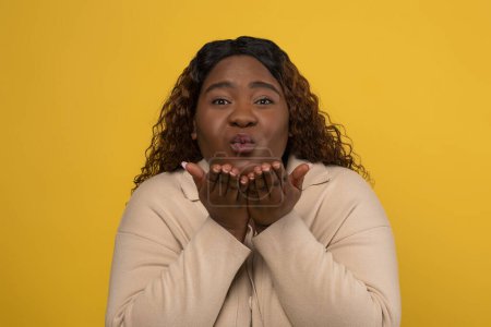Photo for Romantic loving pretty millennial overweight african american lady with long curly hair sending fly kiss at camera, showing her affection, warm feelings, yellow studio background - Royalty Free Image