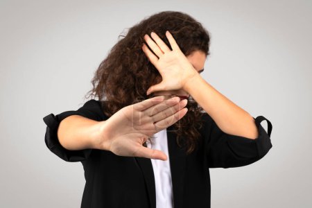 Lockige Geschäftsfrau im Blazer mit entschiedener Stopp-Geste mit der Hand und verdecktem Gesicht, Grenze oder Verweigerung, auf grauem Hintergrund