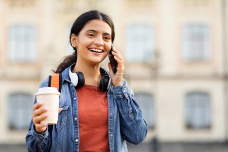 Photo for Portrait of smiling young eastern woman student walking by college campus, talking on phone with friend, holding takeaway coffee, setting meeting after school, copy space - Royalty Free Image
