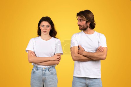 Photo for Serious european young man and woman in plain white t-shirts and blue jeans standing with arms crossed, exhibiting a standoffish attitude, against a vivid yellow background - Royalty Free Image